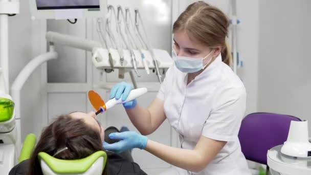 Odontóloga joven en máscara y guantes usando equipo de luz UV dental para endurecimiento de polímeros — Vídeos de Stock