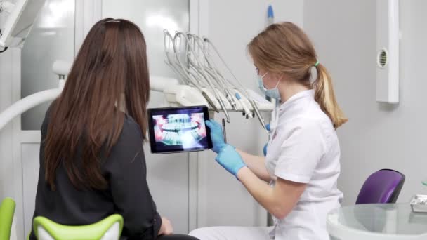 Женщина-дантист показывает рентгеновские зубы на таблетке молодой пациентке. Дантист в халате. — стоковое видео