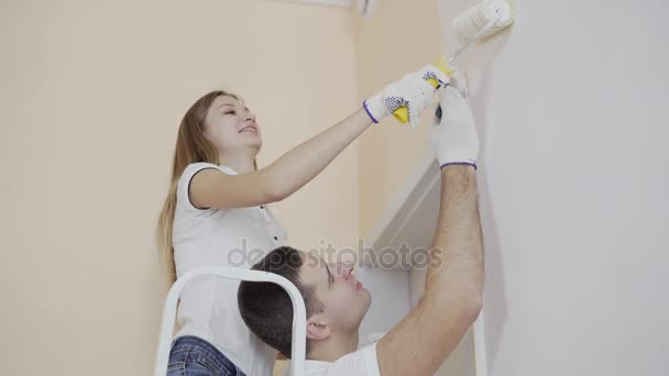 Jeune couple heureux salle de décoration mur de peinture avec rouleau de peinture et pinceau. Femme et homme se tiennent debout sur l'échelle et sourient . — Video
