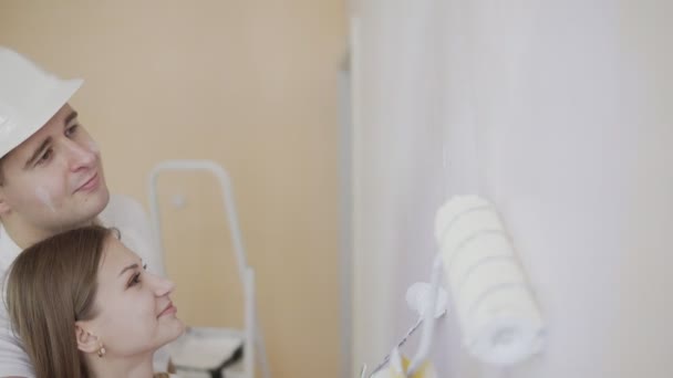 Couple decorating room and painting wall with paint roller in white. Family are preparing their new appartment to move in. Young woman coming up and starting to help — Stock Video