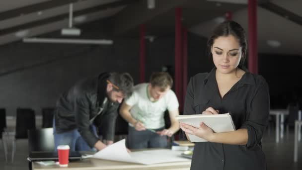 Retrato de uma jovem mulher profissional na moda segurando pastas com papéis e tomando notas em um escritório contemporâneo elegante. Trabalhadores de escritório em segundo plano. Tiros em 4K — Vídeo de Stock