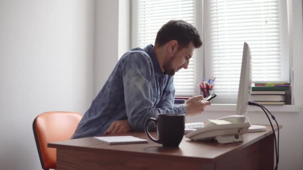 Jonge kantoor werknemer via zijn telefoon op kantoor zitten aan de tafel met de computer, telefoon en cup. Schot in 4k — Stockvideo