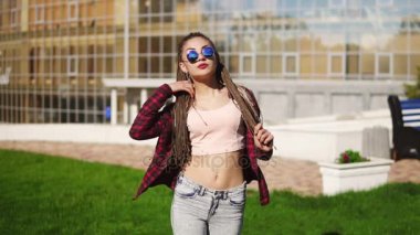 Genç güzel kız dreads bir parkta dans ile. Kot pantolon ve müzik ve güneşli bir gün boyunca dans güneş gözlüğü güzel kadın. Slowmotion atış.