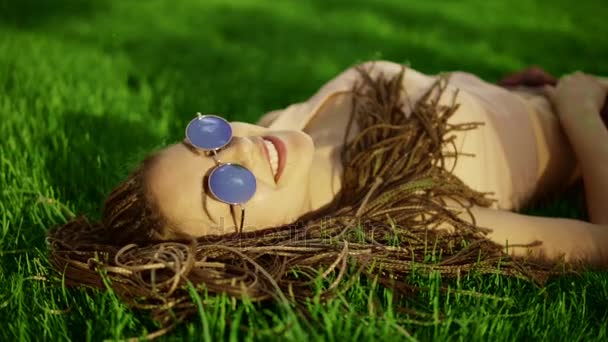 Trendy hipster girl with dreads relaxing on the grass in park. Summer lifestyle portrait of hipster woman laying on the grass, enjoying nice day, wearing bright sunglasses. Slowmotion shot — Stock Video