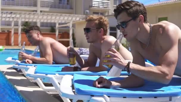Gruppen av unga vänner dricka öl och avkopplande liggande på tränarna vid poolen. Slowmotion skott — Stockvideo
