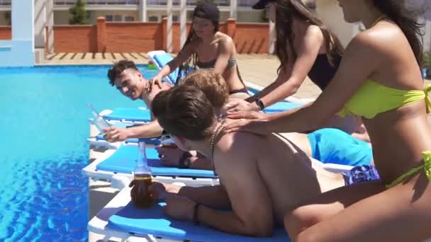 Hombres que reciben masajes de chicas jóvenes atractivas en bikini tumbadas en entrenadores junto a la piscina en una hermosa villa y bebiendo cerveza. Disparo en cámara lenta . — Vídeo de stock