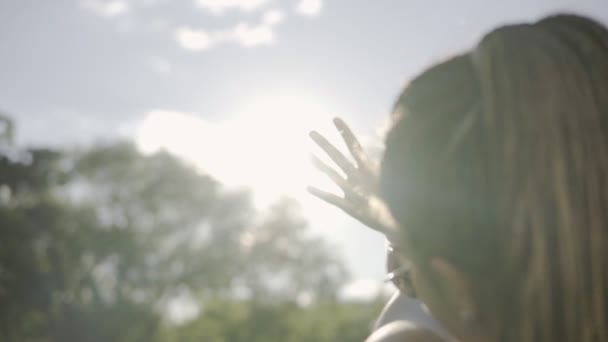 Vista trasera de cerca de una mujer mirando al sol y escondiéndose del sol con su mano. Mujer joven con temores jugando con un rayo de sol. Disparo en cámara lenta . — Vídeos de Stock