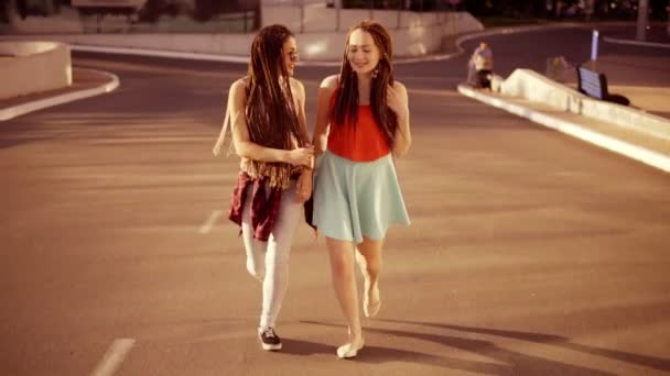 Twee gelukkige vrouwen met dreadlocks op de lege weg lopen en praten in de zomer. Twee meisjes hipster lachen en knuffelen elkaar tijdens een zonnige dag. Tikje schot. — Stockvideo
