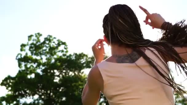 Visão traseira da jovem menina bonita com dreads dançando em um parque. Mulher bonita em óculos de sol ouvindo música e dançando durante um dia ensolarado. Tiro em câmara lenta . — Vídeo de Stock