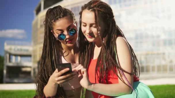Щасливі жінки з страхами сидять на траві в літньому парку і використовують смартфон. Молоді друзі розмовляють і дивляться фотографії, серфінг в Інтернеті на відкритому повітрі розважається. Постріл уповільнення — стокове відео