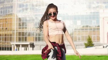 Genç güzel kız dreads bir parkta dans ile. Kot pantolon ve müzik ve güneşli bir gün boyunca dans güneş gözlüğü güzel kadın. Slowmotion atış.