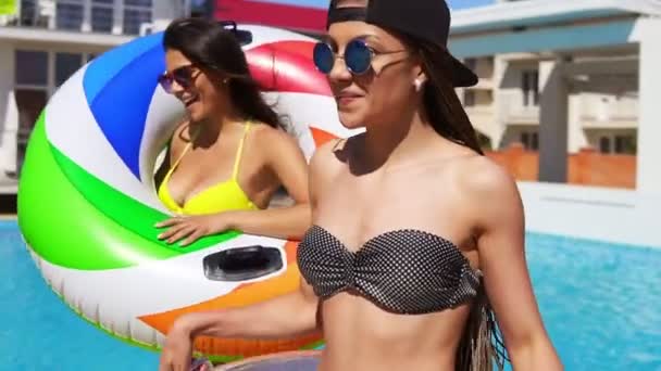 Två attraktiva flickor i solglasögon walking wih uppblåsbara tuber av poolen och leende. Vackra kvinnor som har en poolparty. Slowmotion skott — Stockvideo