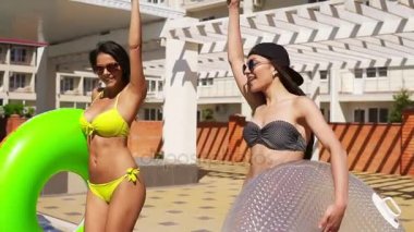 İki çekici kızlar wih şişirilebilir tüp havuz kenarında yürüyüş ve gülümseyen güneş gözlüğü. Güzel kadınlar bir havuz partisi. Slowmotion atış