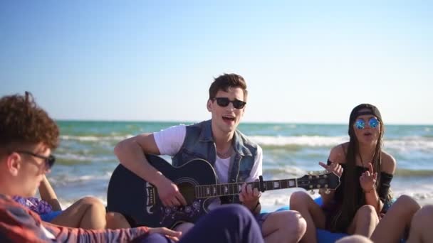 Młody mężczyzna gra git wśród grupy przyjaciół, siedząc na leżaki na plaży i śpiew na letni wieczór. Slowmotion strzał — Wideo stockowe