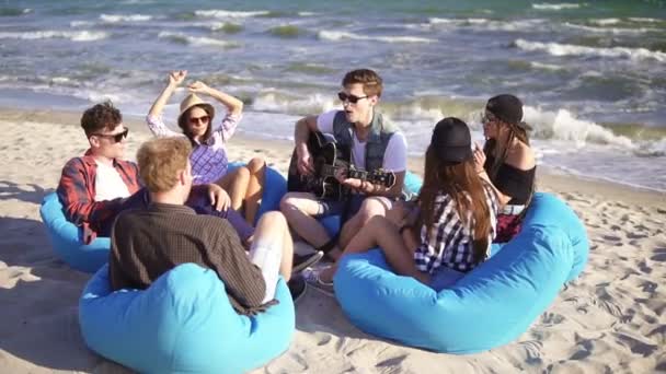 Młody mężczyzna gra git wśród grupy przyjaciół, siedząc na leżaki na plaży i śpiew na letni wieczór. Slowmotion strzał — Wideo stockowe