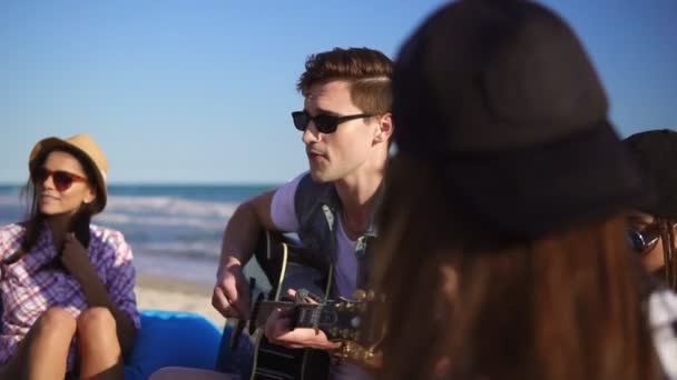 Jovem tocando guitarra entre o grupo de amigos sentados em cadeiras na praia e cantando em uma noite de verão. Tiro em câmara lenta — Vídeo de Stock