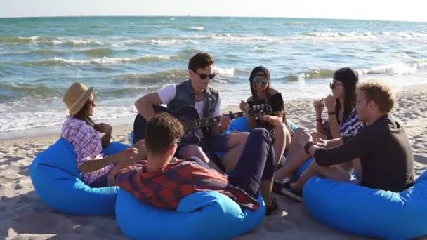 Mladý muž, hrát na kytaru mezi skupinou přátel, sedí na easychairs na pláži a zpívá na letní večer. Slowmotion shot — Stock video