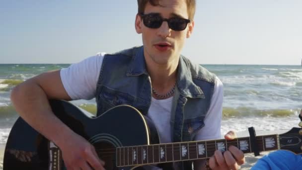 Молодий чоловік грає на гітарі серед групи друзів, сидячи на легких кріслах на пляжі і співаючи в літній вечір. Постріл уповільнення — стокове відео
