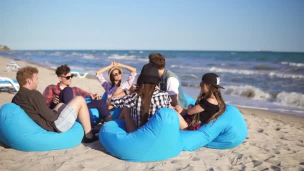 Młody mężczyzna gra git wśród grupy przyjaciół, siedząc na leżaki na plaży i śpiew na letni wieczór. Slowmotion strzał — Wideo stockowe