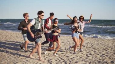 Grup yürüyüş ve birlikte dans gitar çalmak ve bir plaj kenarında sular şarkılar söyleyen genç hipster arkadaşıyla. Slowmotion atış