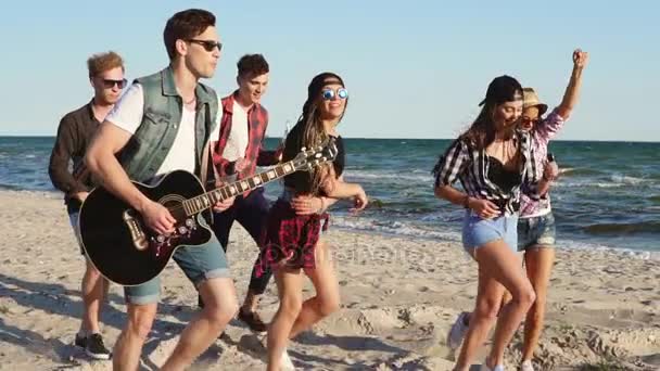 Друзья-хипстеры ходят и танцуют вместе, играют на гитаре и поют песни на пляже на краю воды. Слоумоушн — стоковое видео