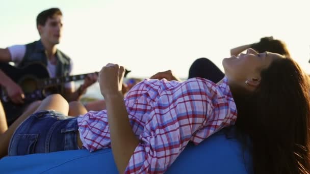 Młoda kobieta na easychair i słuchanie mężczyzna gra na gitarze wśród grupy przyjaciół, siedząc na plaży i śpiewania na letni wieczór podczas zachodu słońca. Slowmotion strzał — Wideo stockowe