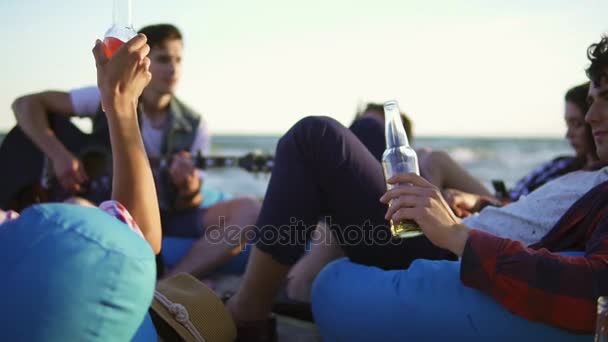 Grupo de amigos tomando cócteles y cerveza y haciendo vítores sentados en sillas cómodas en la playa y escuchando a un amigo tocando la guitarra en una noche de verano durante una puesta de sol. Disparo en cámara lenta — Vídeos de Stock