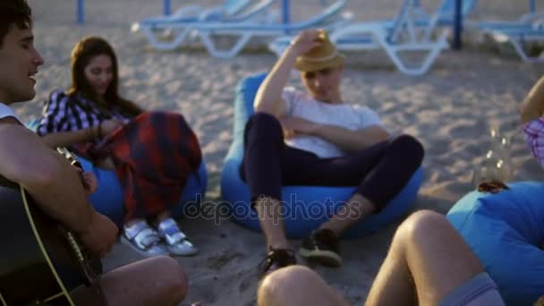 Genç adam sahilde easychairs üzerinde oturan ve bir yaz akşamı bir gün batımı sırasında şarkı arkadaş grubu arasında gitar çalıyor. Slowmotion atış — Stok video