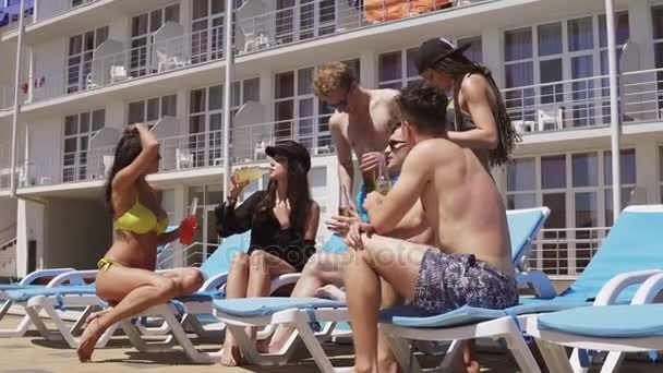 Grupo de hermosos jóvenes amigos en trajes de baño bebiendo cócteles, hablando y divirtiéndose sentados junto a la piscina. Disparo en 4k — Vídeo de stock