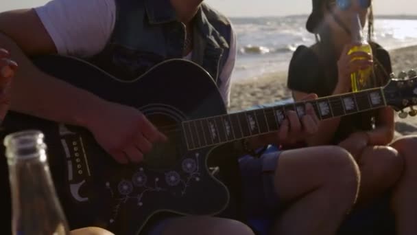 Jonge happy vrienden drinken bier, zittend op de easychairs op het strand en het luisteren naar een vriend gitaarspelen op een zomeravond. Schot in 4k — Stockvideo
