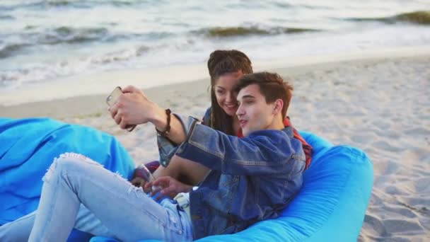 Пара розважається і приймає селфі, лежачи на простих стільцях з ковдрою на пляжі. Постріл в 4k — стокове відео