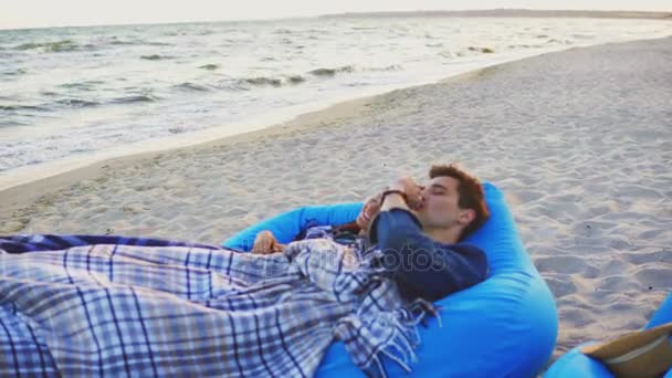 Paar, das auf Liegestühlen am Strand liegt und mit einer Decke bedeckt ist. Mann, der elektronische Zigarette raucht. Dampf ab. Schuss in 4k — Stockvideo