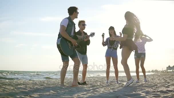 Fiesta de verano en la playa. Jóvenes amigos bebiendo cócteles, bailando en el círculo, tocando la guitarra, cantando canciones y aplaudiendo en una playa en el borde del agua durante la puesta del sol. Disparo en cámara lenta — Vídeos de Stock