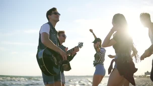 Sommerfest am Strand. Junge Freunde trinken Cocktails, tanzen im Zirkel, spielen Gitarre, singen Lieder und klatschen während des Sonnenuntergangs am Strand am Rande des Wassers. Zeitlupenschuss — Stockvideo