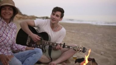 Genç adam ateşle birlikte arkadaşlarıyla sahilde oturup gitar çalıyor. Kız arkadaşı onun için ızgara sosis getiriyor. Slowmotion atış