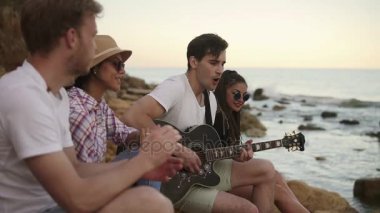 Seashore tarafından kayaların üzerinde oturan ve gitar ve şarkı oynayan genç hipster arkadaş grubu. Slowmotion atış
