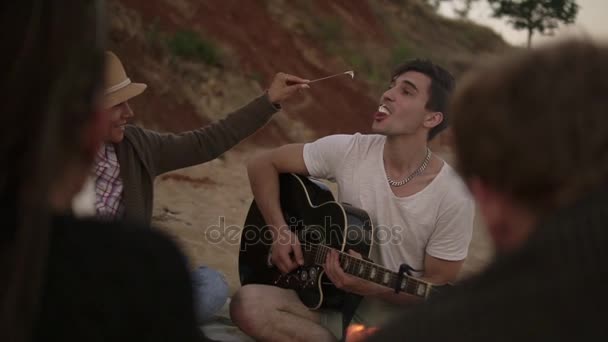 Romantisches junges Paar, das mit Freunden am Feuer am Strand sitzt. Der Mann spielt eine Gitarre und die Frau reicht ihm einen Sumpf-Malve. Zeitlupenschuss — Stockvideo