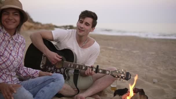 Młody człowiek jest gra na gitarze przez ogień, siedząc na plaży wraz z przyjaciółmi. Jego dziewczyna, przynosząc Kiełbasa z grilla dla niego. Slowmotion strzał — Wideo stockowe