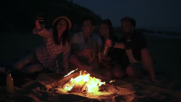 Picnic of young people with bonfire on the beach in the evening. Cheerful friends taking pictures on the phone. Slowmotion shot — Stock Video