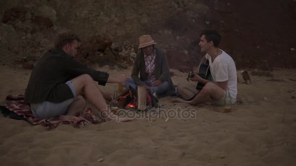 Giovani amici seduti accanto al fuoco sulla spiaggia la sera, grigliando salsicce, bevendo birra e suonando la chitarra. Colpo di rallentamento — Video Stock