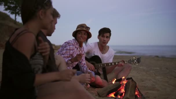 Picnic di giovani con falò sulla spiaggia la sera. Amici allegri che cantano canzoni e suonano la chitarra. Colpo di rallentamento — Video Stock