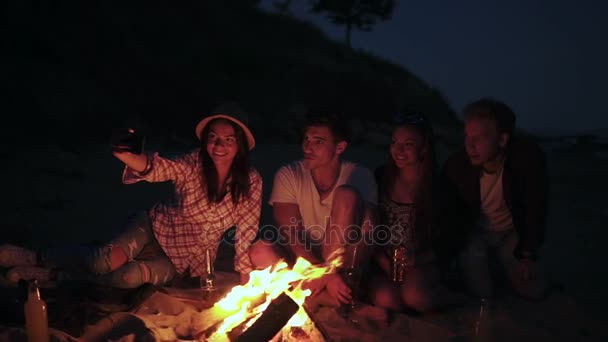 Picnic di giovani con falò sulla spiaggia la sera. Amici allegri che scattano foto al telefono. Colpo di rallentamento — Video Stock