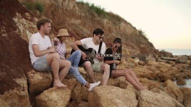 Grup şarkıları şarkı ve dans seashore tarafından kayaların üzerinde oturan ve gitar, çalmak genç hipster arkadaşıyla. Slowmotion atış