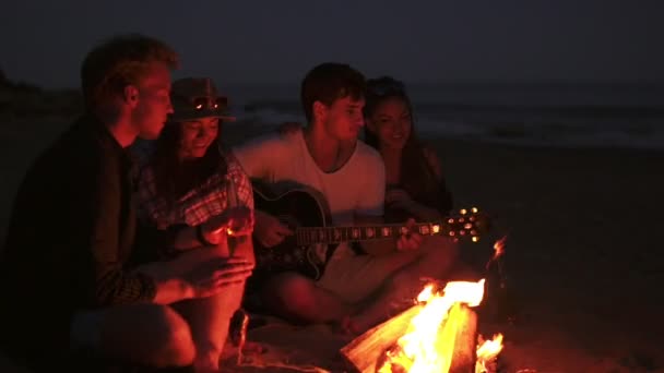 Piknik młodych ludzi z ogniskiem na plaży w godzinach wieczornych. Wesoły znajomi, śpiewając piosenki i gra na gitarze. Slowmotion strzał — Wideo stockowe