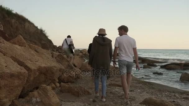 Ομάδα των νέων και χαρούμενο περπάτημα από την παραλία και να κάθονται πάνω στα βράχια το βράδυ, παίζοντας κιθάρα και τραγουδώντας τραγούδια. Slowmotion βολή — Αρχείο Βίντεο