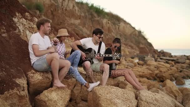 Groupe de jeunes amis hipster assis sur les rochers au bord de la mer et jouant de la guitare, chantant des chansons et dansant. Coup de ralenti — Video