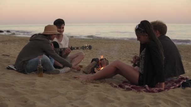 Jóvenes amigos alegres sentados junto al fuego en la playa por la noche, bebiendo cerveza y tocando la guitarra. Disparo en 4k — Vídeo de stock