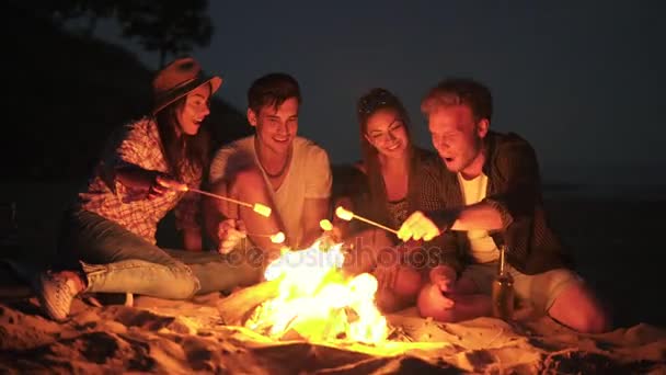 Jóvenes amigos alegres sentados junto al fuego en la playa por la noche, cocinando malvavisco en palos juntos. Disparo en 4k — Vídeos de Stock
