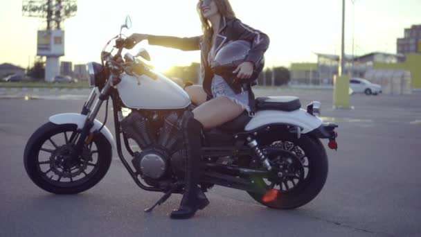 Linda motociclista en pantalones cortos sentada en el asiento de la motocicleta y sosteniendo su casco. Primer plano de la joven mujer rizada sexy en gafas de sol en el helicóptero durante la puesta del sol — Vídeos de Stock