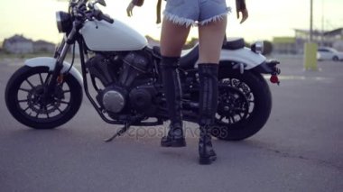 Arkadan geliyor ve motosiklet üzerinde getting şortlu kadın bir bisikletçinin görünüşü. Genç seksi kıvırcık kadın helikopter üzerinde güneş gözlüğü